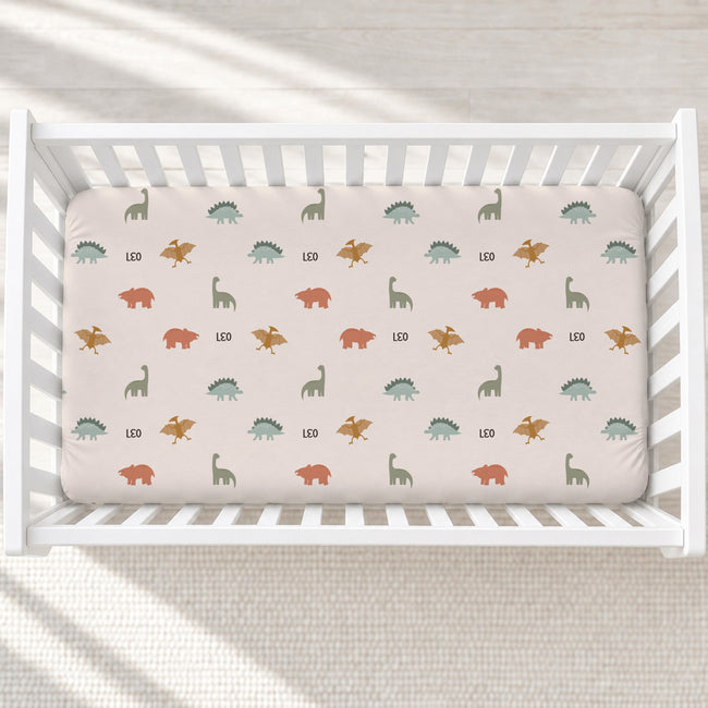 A fitted crib sheet with a dinosaur pattern in muted green, orange, and brown on a light blush background, shown inside a white wooden crib.