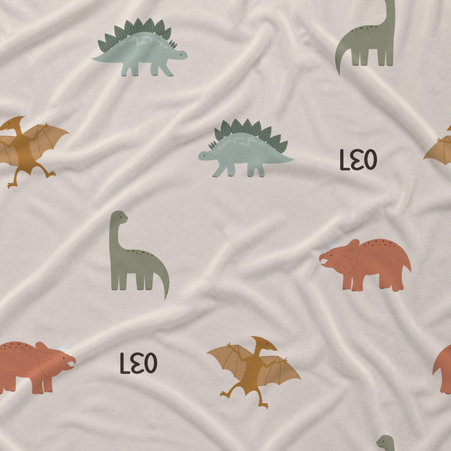 A close-up of a soft fabric surface with an allover dinosaur print on a light warm beige background. The pattern includes sage green long-neck dinosaurs, muted blue-green Stegosaurus-like dinosaurs, and terracotta-brown dinosaurs, with the word 'LEO' printed in black.