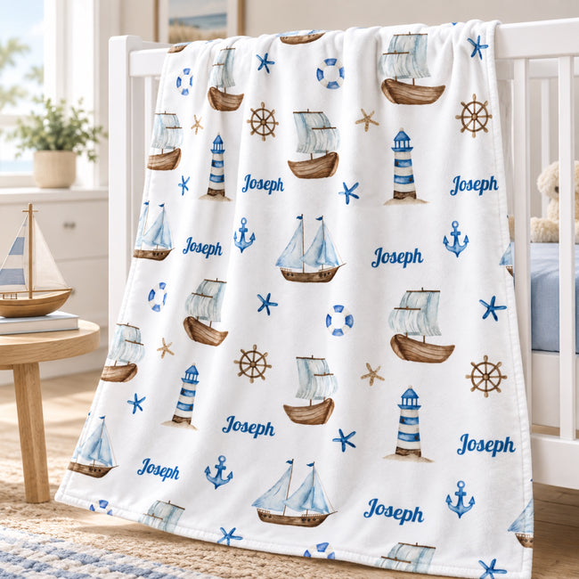 A white baby blanket with a nautical theme, featuring blue and brown illustrations of sailboats, lighthouses, anchors, and stars, along with the name 'Joseph' printed in blue. The blanket is draped over a white wooden crib in a bright, modern nursery setting.