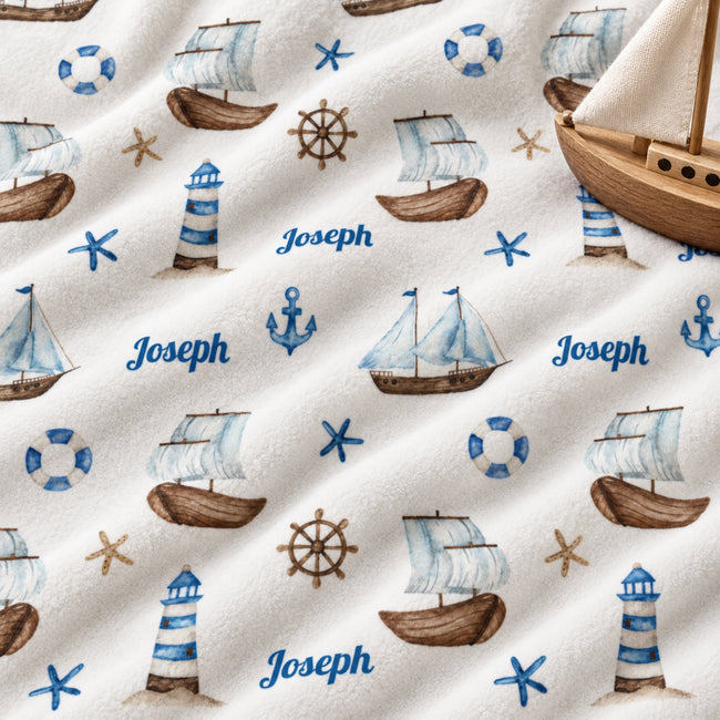 Personalized baby fabric with a nautical theme, featuring blue and brown watercolor-style illustrations of sailboats, lighthouses, anchors, life buoys, starfish, and ship's wheels, with the name 'Joseph' printed in blue script. A wooden toy boat is placed on top of the fabric.