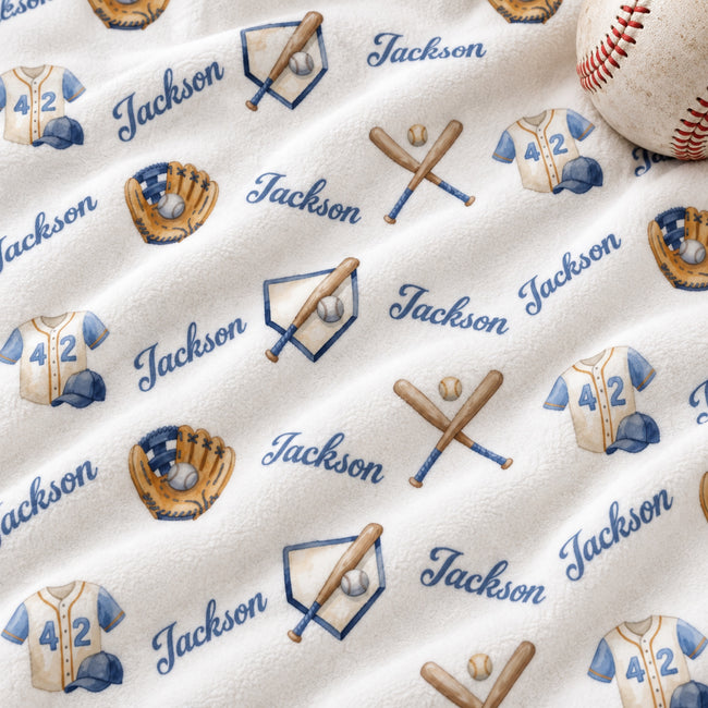 A close-up of a personalized fleece blanket featuring a baseball theme. The blanket has a cream-colored background with a repeating pattern of navy blue script text reading 'Jackson', blue and navy baseball uniforms with the number '42', crossed wooden bats, baseballs, and baseball gloves.