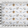 A close-up of a white fitted sheet with a repeating baseball-themed pattern featuring the name 'Jackson' in blue script, crossed bats, baseball gloves, and jerseys, shown inside a white crib.