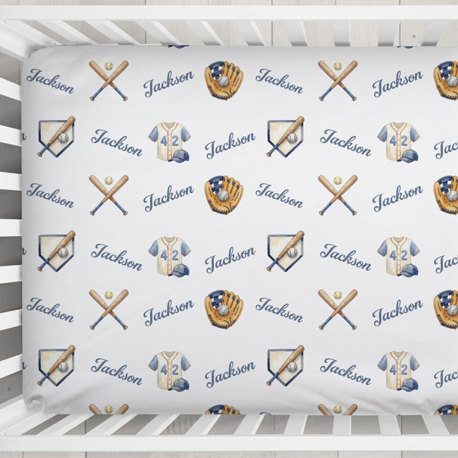 A close-up of a white fitted sheet with a repeating baseball-themed pattern featuring the name 'Jackson' in blue script, crossed bats, baseball gloves, and jerseys, shown inside a white crib.