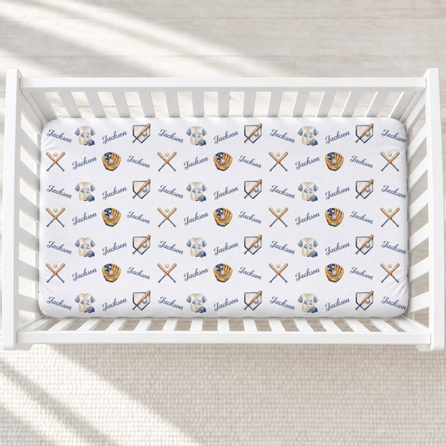 A white crib mattress fitted sheet with a repeating pattern of navy blue 'Jackson' text, baseball caps, baseball gloves, and crossed bats, shown inside a white wooden crib.
