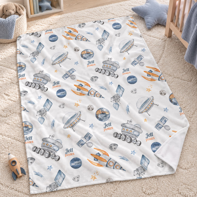 Personalized Rocket Baby Blanket. Rockets, satellites, planets, and space rovers surround a custom name in watercolor blue, grey, and orange.