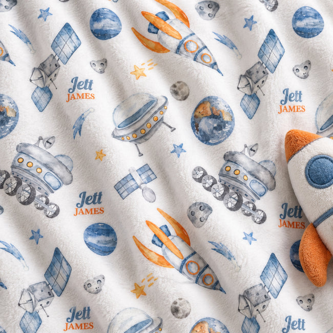 A close-up of a plush white blanket or fabric featuring a repeating space-themed print with blue planets, gray satellites, orange rockets, stars, and the personalized name 'Jett James' repeated in blue and orange.