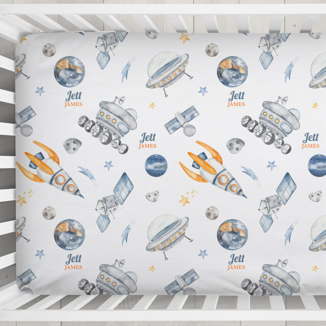A crib mattress fitted sheet with a personalized space-themed print featuring watercolor-style rockets, satellites, planets, stars, and the text 'Jett James'.
