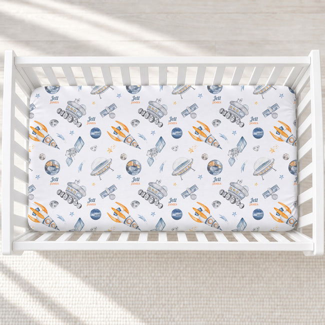 A fitted crib mattress with a white background and a colorful space-themed print featuring rockets, satellites, rockets, and planets. The crib is shown from a top-down view on a light-colored floor with soft shadows.