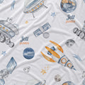 A white fabric product, likely a baby blanket or quilt, featuring a watercolor space-themed print with rockets, satellites, planets, stars, and the personalized name 'Jett James' in blue and orange text.