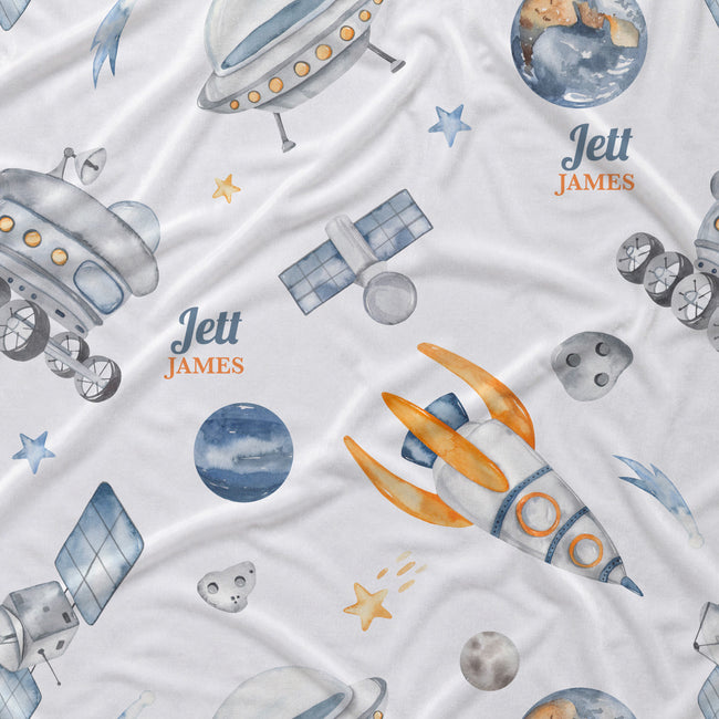 A white fabric product, likely a baby blanket or quilt, featuring a watercolor space-themed print with rockets, satellites, planets, stars, and the personalized name 'Jett James' in blue and orange text.
