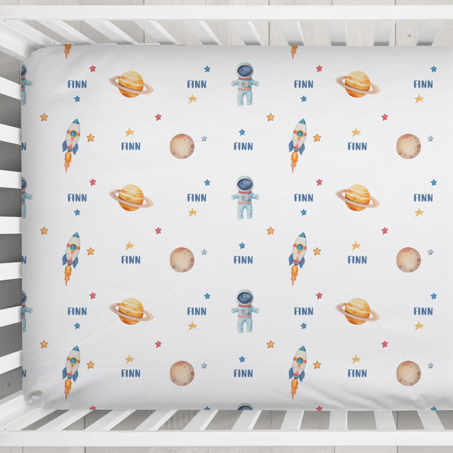 A white fitted crib sheet with a repeating space-themed print featuring astronauts, rockets, planets, stars, and the name 'FINN' in blue, shown inside a white wooden crib.