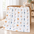 A white baby blanket with a colorful space-themed print featuring astronauts, rockets, planets, and stars, draped over a woven basket with a teddy bear on a wooden floor.