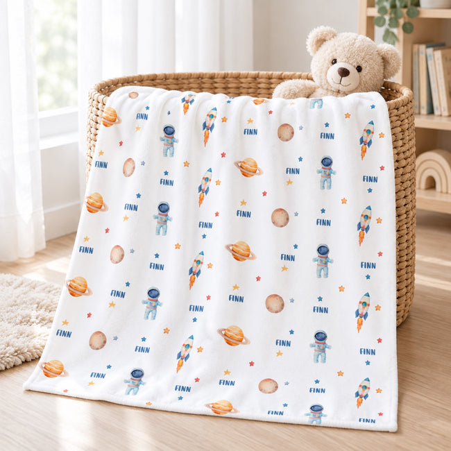A white baby blanket with a colorful space-themed print featuring astronauts, rockets, planets, and stars, draped over a woven basket with a teddy bear on a wooden floor.