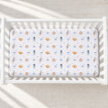A fitted crib mattress with a white background and a repeating space-themed print featuring astronauts, rockets, planets, and stars in blue, orange, and brown, shown inside a white wooden crib.