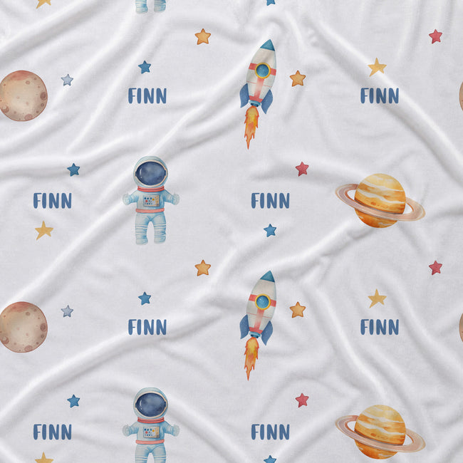 A white fabric with a repeating space-themed print featuring astronauts, rockets, planets, stars, and the name 'FINN' in blue text.