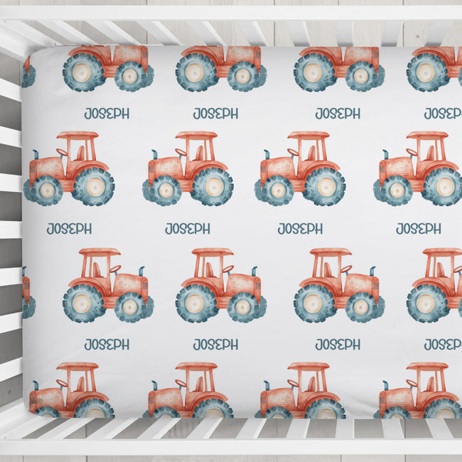 A crib sheet featuring a repeating pattern of red-orange watercolor tractors with blue tires and the name 'JOSEPH' printed underneath each tractor, shown inside a white wooden crib.