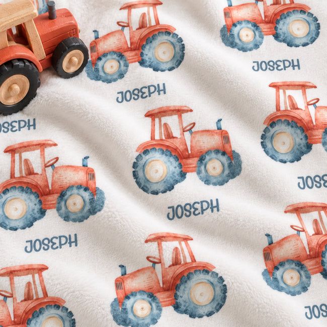 A close-up of a white fleece blanket printed with a repeating pattern of red-orange watercolor tractors with blue tires and the name 'JOSEPH' printed below each tractor.