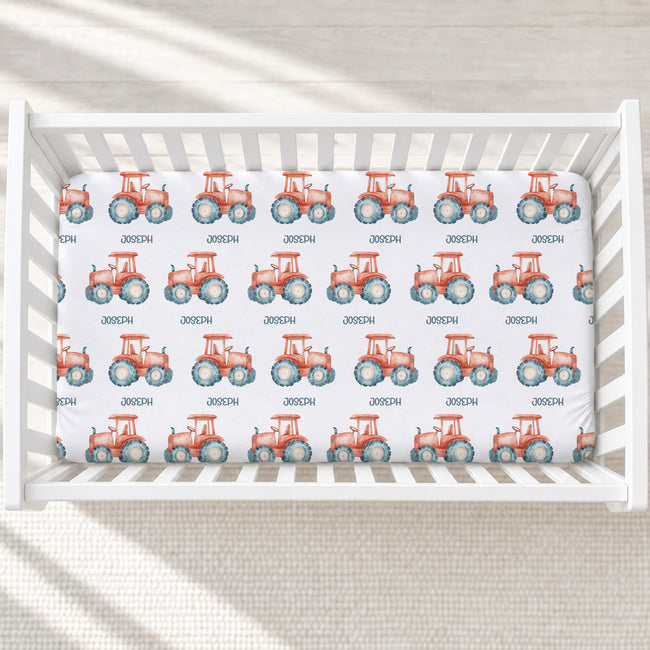 A white crib mattress fitted sheet printed with a repeating pattern of orange-red tractors with blue wheels and small black text, shown inside a white crib on a light neutral floor.