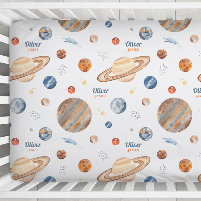 A personalized baby crib sheet with a space-themed print featuring planets, stars, and the names 'Oliver' and 'JAMES' in blue and orange, displayed inside a white wooden crib.