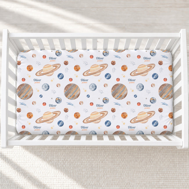 A white crib mattress fitted with a space-themed sheet featuring colorful illustrations of planets, stars, and the name 'Oliver' printed repeatedly, shown in a softly lit nursery setting.