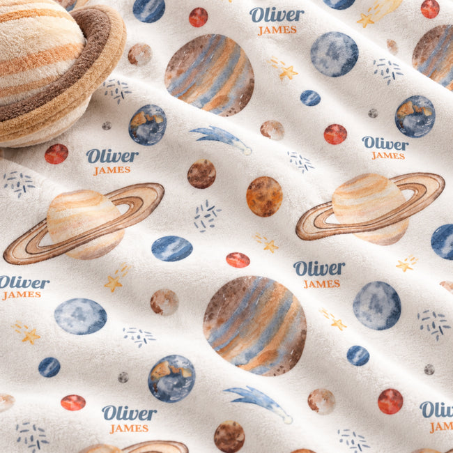 Close-up of a soft fabric product printed with planets, stars, and repeated 'Oliver James' text on a cream background. A small plush planet toy is partially visible in the upper left corner.