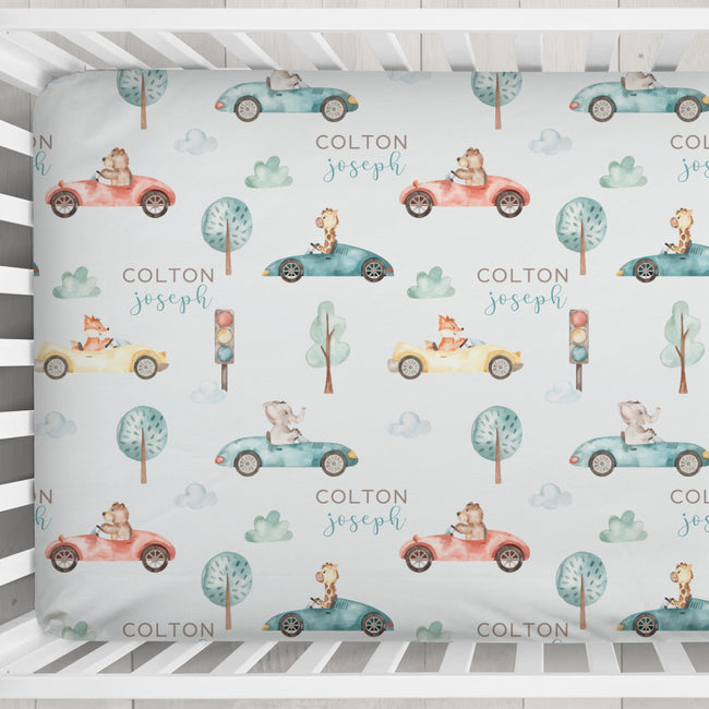 A personalized baby crib bumper sticker with a repeating pattern of cute cartoon animals in vintage cars, trees, and the names 'COLTON joseph'. The bumper sticker is shown fitted inside a white wooden crib.