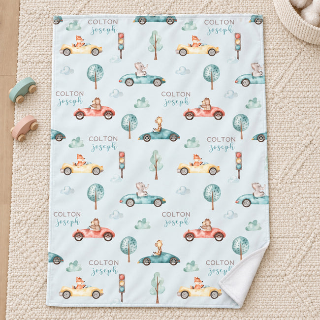 Personalized Animal Race Car Baby Blanket. Friendly animals drive colorful race cars among trees, clouds, and traffic lights around a custom name.