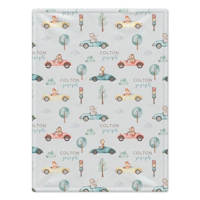 Personalized Animal Race Car Baby Blanket. Friendly animals drive colorful race cars among trees, clouds, and traffic lights around a custom name.