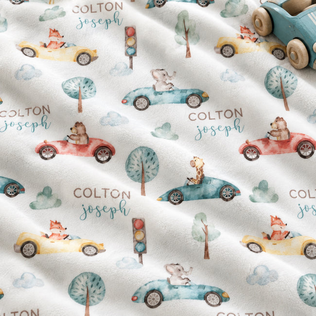 Personalized Animal Race Car Baby Blanket. Friendly animals drive colorful race cars among trees, clouds, and traffic lights around a custom name.