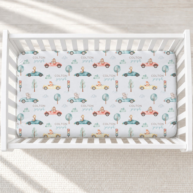 A white wooden crib with a fitted sheet printed with a whimsical pattern of vintage cars, trees, and the name 'COLTON Joseph' repeated across a white background.
