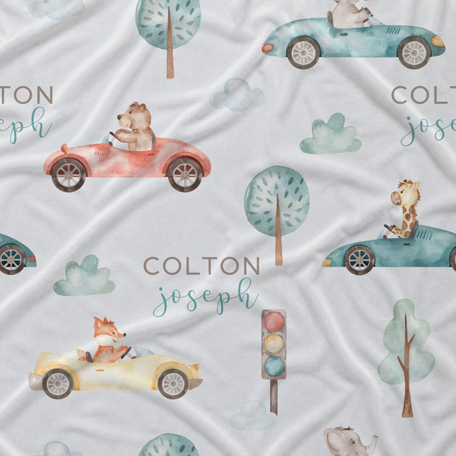 Personalized baby bedding fabric featuring a whimsical watercolor pattern of animals driving colorful cars on a white background, with the custom name 'COLTON joseph' printed in teal and brown.