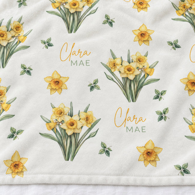 A close-up of a white fabric featuring a repeating pattern of yellow daffodils, small green leaves, and the personalized name 'Clara MAE' in gold and green lettering.