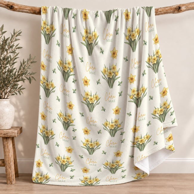 Ivory blanket with yellow daffodil clusters, scattered green leaves, and the name Clara Mae in gold and sage green lettering.