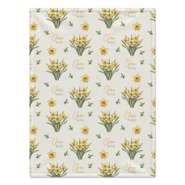 Ivory blanket with yellow daffodil clusters, scattered green leaves, and the name Clara Mae in gold and sage green lettering.