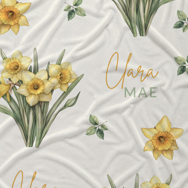 Ivory fabric with yellow daffodil clusters, scattered green leaves, and the name Clara Mae in gold and sage green lettering.