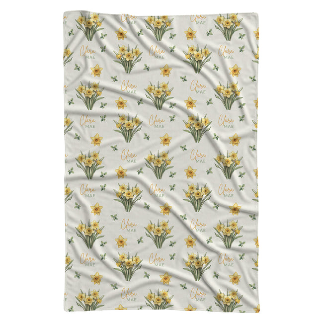 Ivory swaddle blanket with yellow daffodil clusters, green leaves, and the name Clara Mae in gold and sage green lettering.