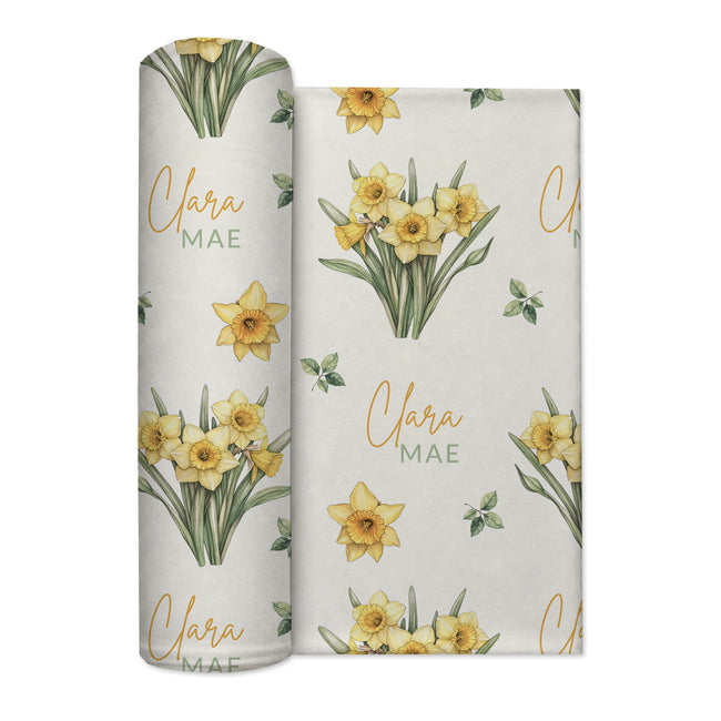 Ivory swaddle blanket with yellow daffodil clusters, green leaves, and the name Clara Mae in gold and sage green lettering.