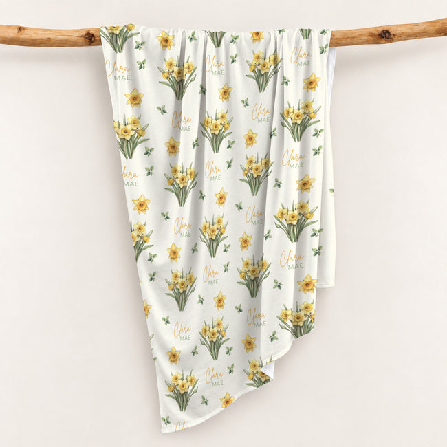 Ivory swaddle blanket with yellow daffodil clusters, green leaves, and the name Clara Mae in gold and sage green lettering.