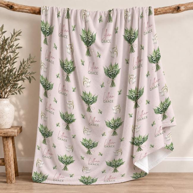 Pale blush blanket with white lily of the valley bouquets, green leaves, and the name Lillian Grace in rose and green lettering.