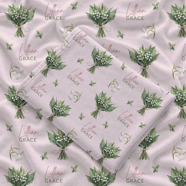 Pale blush swaddle blanket with white lily of the valley bouquets, green leaves, and the name Lillian Grace in rose and green.