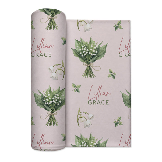 Pale blush swaddle blanket with white lily of the valley bouquets, green leaves, and the name Lillian Grace in rose and green.