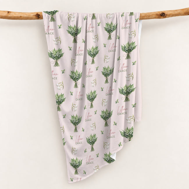 Pale blush swaddle blanket with white lily of the valley bouquets, green leaves, and the name Lillian Grace in rose and green.