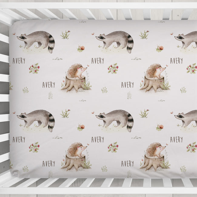 A crib fitted sheet with a repeating woodland print featuring raccoons, hedgehogs, flowers, and the name 'Avery' in a nursery setting.