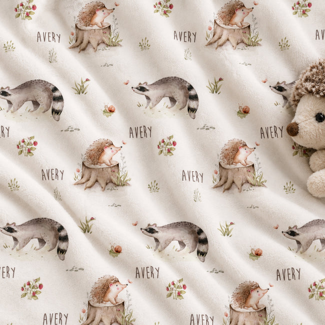 Personalized Woodland Animal Baby Blanket. Sweet hedgehogs, raccoons, snails, and meadow plants surround a custom name on a soft neutral background.