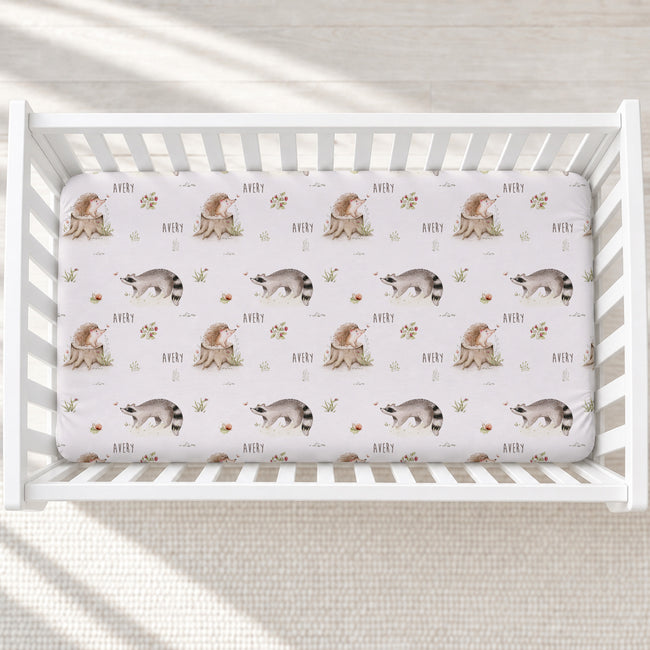 A fitted crib mattress with a repeating pattern of raccoons, squirrels, and the name 'AVERY' in a soft watercolor style, shown inside a white wooden crib on a light neutral floor.