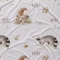 A white fabric with a whimsical woodland print featuring a hedgehog on a tree stump, a raccoon, a snail, small plants, and the name 'AVERY' repeated in dark gray text.