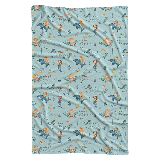A rectangular baby blanket with a repeating mermaid and dolphin pattern on a light blue background, featuring the name 'Anna' printed in gold script.