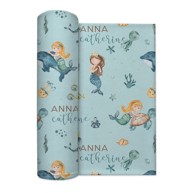 A rolled personalized swaddle blanket featuring a light blue mermaid-themed pattern with illustrations of mermaids, sea turtles, octopus, and starfish, along with the custom text 'ANNA catherine' in gold and dark blue script.