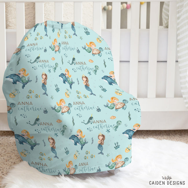 A custom car seat cover with a light blue mermaid pattern, featuring the name 'Anna Catherine' in gold script, displayed on a white car seat in a nursery setting.