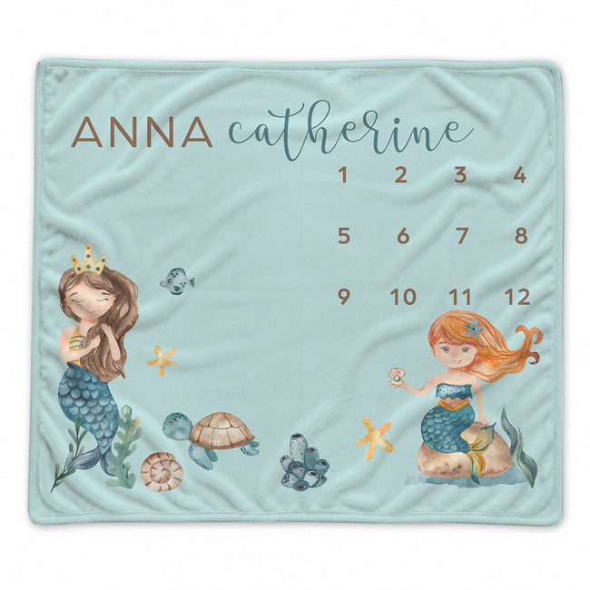 A personalized baby blanket featuring a light blue background with watercolor illustrations of two mermaids, a turtle, starfish, and a calendar grid with the names 'ANNA' and 'catherine' at the top.